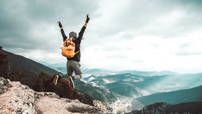 Person mit Rucksack springt auf einem Berg mit Blick auf eine weite Landschaft.
