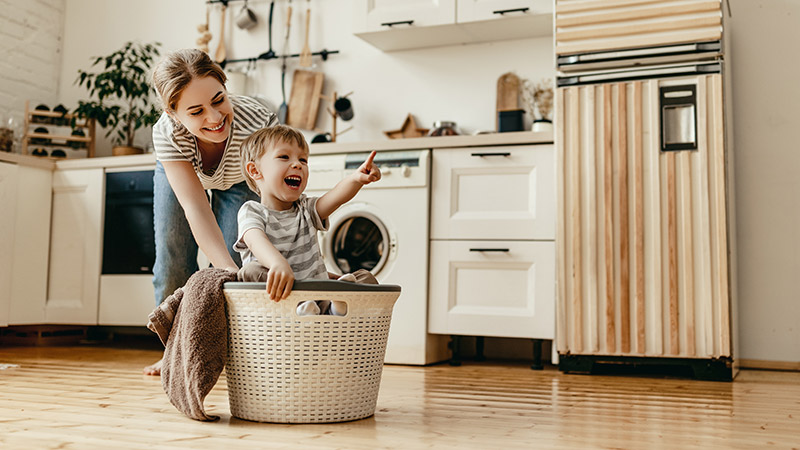 Eine Mutter schiebt ihren lachenden Sohn in einem Wäschekorb durch die Küche.